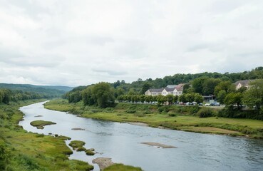 Obraz premium Serene river meanders through green valley under soft clouds. Gentle water reflects trees, sky along grassy banks. Distant village with houses, cars adds to peaceful afternoon scenery. Harmony with