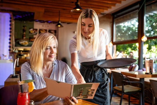 Senior woman sitting in restaurant for lunch, looking in menu, Hospitable waitress help to customer what to choose. Good service in restaurant and helpful employee. 