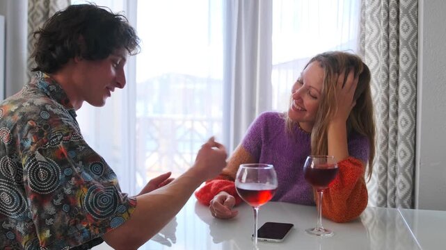 The couple spends time in a cottage, drinking red wine by a large window. The man gently holds the woman hand as she leans on her elbow, smiling and watching him with a fond, flirty smile.