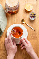 Hands holding cup of herbal tea with lemon on a wooden table with teapot and napkin.