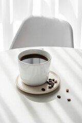 Freshly brewed espresso coffee in a cup on a white table near window with chair, coffee beans