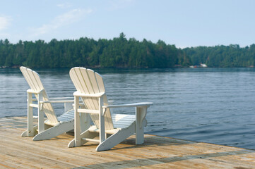 White Adirondack chairs perched on a dock overlook a forest-lined lake in Ontario, basking in the warm glow of early morning sun. The scene captures the essence of a serene summer retreat.