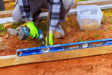 Contractor in gloves drills into wood while using level to ensure accuracy in backyard construction project.