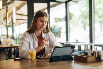 Eating, digital tablet is on the table. Young woman is in the cafe at daytime