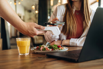 Laptop, salad and orange juice. Woman is paying for meal by wireless payment in the cafe restaurant
