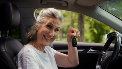 Happy smiling elderly senior mature Caucasian woman female lady driver car owner buyer rental service client looking at camera friendly smile showing automobile keys buying rent vehicle inside auto