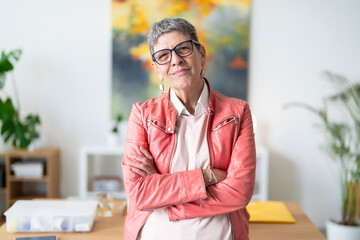 Middle aged woman in glasses and pink leather jacket crossing arms and looking at camera while standing in modern office