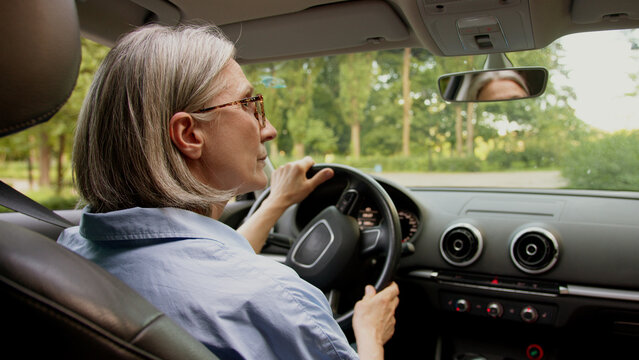Back view Caucasian mature elderly business woman aged female businesswoman in glasses taxi driver hold steering wheel steer auto inside automobile rearview mirror carefully driving machine attention
