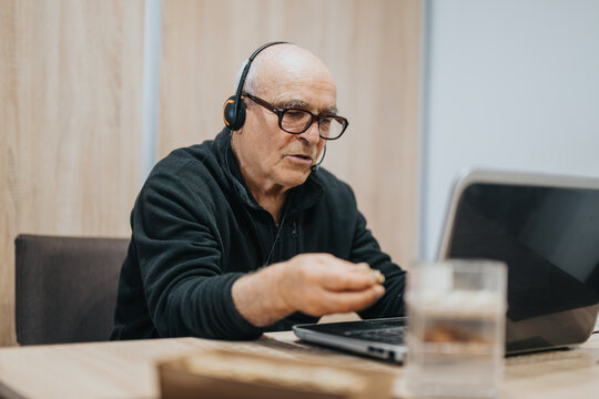 Senior man with glasses wearing headphones using a laptop, participating in an online discussion or virtual meeting, creating a sense of focus, technology use, and digital communication at home.