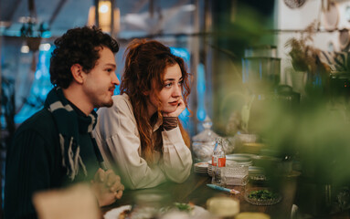 A couple seated at a bar surrounded by warm lighting and decor, engaging attentively together in a lively ambiance. Bottles and plates suggest a relaxed and informal dining experience.