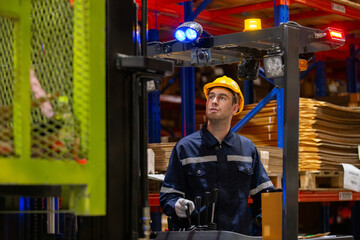 Industrial worker operating forklift in warehouse while wearing safety gear and uniform. Focused male employee managing inventory logistics and lifting materials in storage facility.