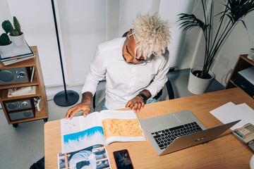 From above direct flash shot of Middle Eastern man in white shirt and blond dreadlocks reading article and examining pictures in magazine while sitting and working on design project in workplace.