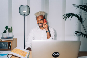 Direct flash shot of happy Middle Eastern designer in white shirt and blond dreadlocks smiling and having smartphone conversation while sitting at desk with magazine and laptop in workspace.