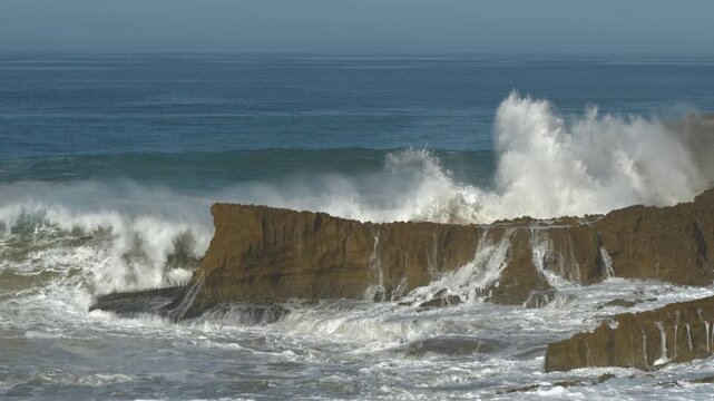 SLOW MOTION: Huge wave crashes against a cliffside, sending enormous amount of white water and foam high into the air. Water cascades down the face of a scenic cliff and spills over the rocks below.
