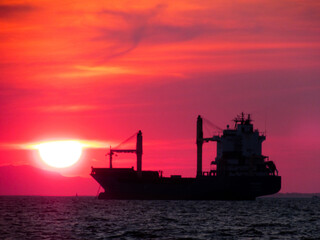 Fototapeta premium Cargo ship sailing at sea during a stunning sunset, silhouette view with warm colors in the sky.