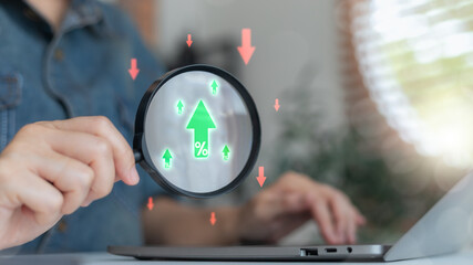 Close-up of hand holding magnifying glass focusing on green upward arrow with percentage symbol...