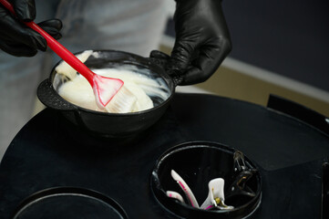 Close-up of hands wearing black gloves mixing creamy white hair dye in a black bowl with a red...