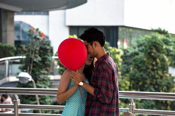 Romantic young couple sharing intimate moment behind red balloon on outdoor skywalk, surrounded by...