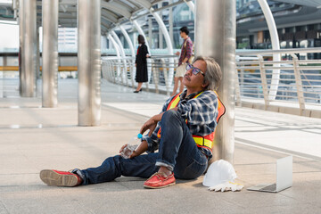 Exhausted Southeast Asian construction worker in safety vest and helmet sitting on city walkway,...
