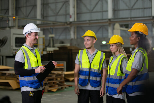 Group of diverse industrial workers in safety vests and helmets listening to supervisor during briefing at factory, showing teamwork, training, discipline and communication in workplace safety.