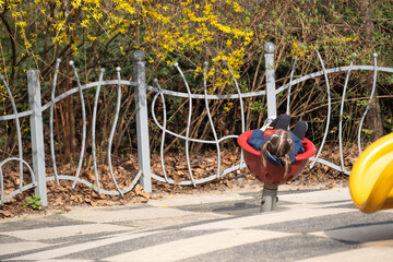 A girl with pigtails sits in a red spinning playground seat, surrounded by dry leaves and flowering...