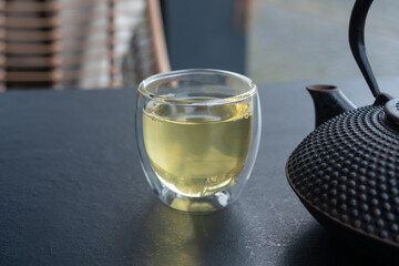 Glass cup with green tea and cast iron teapot on dark table, double wall glass