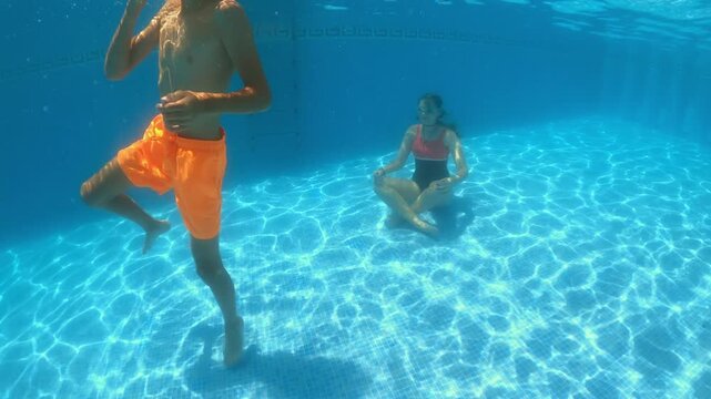 Young boy in orange shorts dancing playfully underwater beside teenage girl meditating in one-piece swimsuit at swimming pool bottom