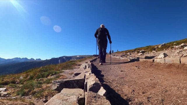 A man hikes uphill on a rocky mountain trail using trekking poles. Hiking and mountain idea. Outdoor adventure and exercise concept.