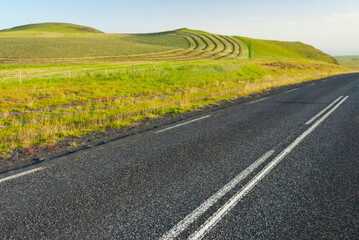 Fototapeta premium A quiet country road runs past green hills and patterned fields, capturing the peaceful rhythm of rural life and the beauty of open landscapes in soft evening light.