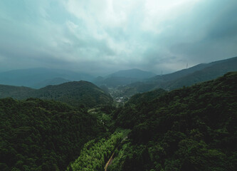 Naklejka premium A vast, misty mountain range shrouded in clouds, with dense green forests and a winding road. A small village nestled in the valley below. 