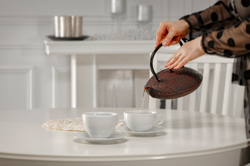 Woman’s hand pouring hot tea from ornate decorative teapot into cup in bright yet warm and cozy home interior. Copy space for product placement.