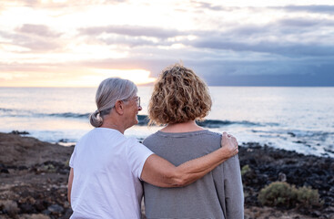 Back view of a romantic senior couple of women embracing on the seashore at sunset light looking at...