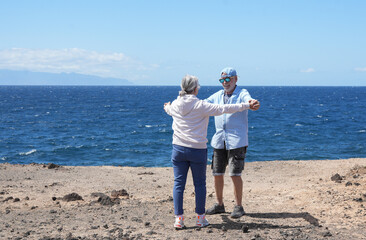 A joyful elderly couple dances while strolling by the sea under a blue sky. Two cheerful retirees, dressed casually, radiate love and happiness, enjoying each other's company.