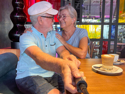 Joyful senior couple sharing a warm moment over coffee in a modern café. The man holds a selfie stick while the woman looks at him lovingly