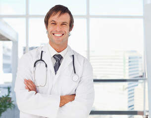 Mid-adult male doctor standing in lobby wearing coat and tie, holding stethoscope while smiling