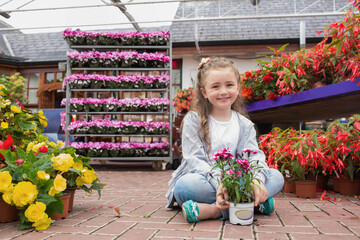 Female child sitting cross-legged holding white pot with pink blossoms on brick floor in greenhouse