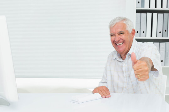 Senior man smiling and extending arm for thumbs-up sitting at office desk with monitor, copy space