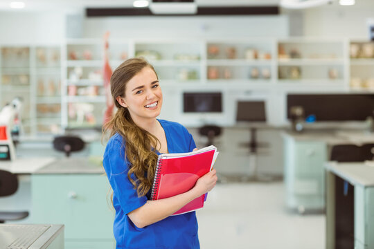 Smiling female student wearing blue scrubs and holding red spiral notebook in science lab classroom