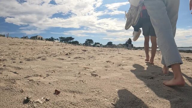 Tracking shot of two people walking barefoot on sandy beach carrying shoes on a sunny day. Summer vacation and beach walk idea. Coastal relaxation and outdoor lifestyle concept.