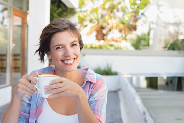 Woman in twenties holding white ceramic coffee mug at outdoor café terrace wearing plaid shirt
