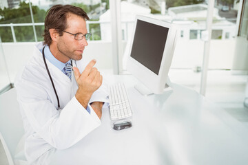 Doctor wearing white coat and stethoscope pointing at monitor while sitting at desk, copy space