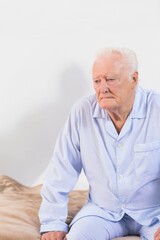 Senior man leaning forward on bed edge in pajamas near beige blanket and mattress, copy space