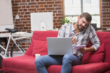 Man calling on smartphone while writing in notepad on red sofa near bicycle in loft office