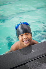African American child boy leaning on pool deck at turquoise pool wearing black cap, blue goggles