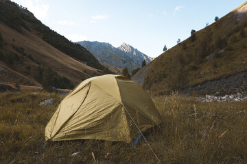Yellow camping tent pitched in a wild alpine valley, surrounded by dry summer grass and mountains in the background