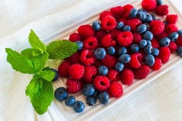Close-up of ripe red raspberries and blueberries arranged on a white ceramic plate with fresh mint...