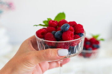 Woman’s hand holding a small glass bowl filled with raspberries, blueberries, and mint. Soft light and white background create a clean minimalistic look.