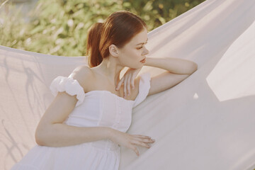 A serene young woman in a white dress relaxing in a hammock, surrounded by lush greenery, conveying...