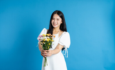 Smiling woman in a white dress holding a bunch of colorful flowers against a solid blue background