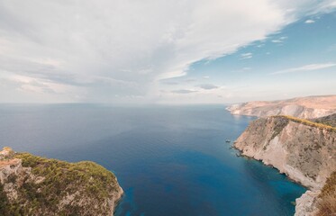From a high vantage point, sheer cliffs plunge dramatically into the Aegean Sea. The deep blue water contrasts beautifully with the rocky landscape on a cloudy afternoon in Zakynthos